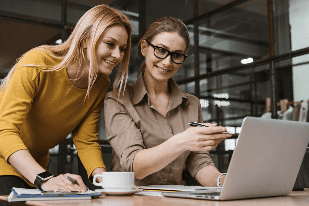Duas mulheres trabalhando juntas em um ambiente de escritório moderno, uma delas aponta para o laptop enquanto a outra faz anotações. O foco está em colaboração e produtividade.