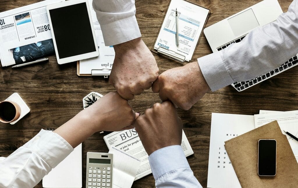 Cinco mãos se encontrando em um gesto de união, simbolizando trabalho em equipe e colaboração em um ambiente de negócios. Escritório com materiais de trabalho ao fundo.
