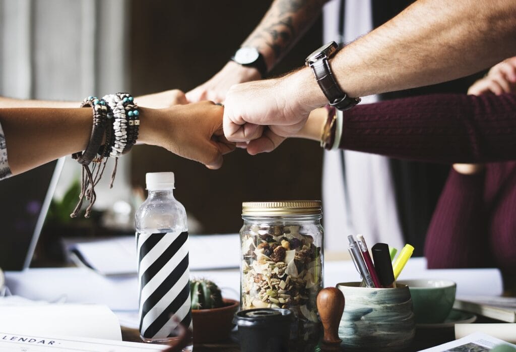 Grupo de pessoas fazendo um gesto de empoderamento com as mãos unidas, representando trabalho em equipe e colaboração em um ambiente de trabalho descontraído.