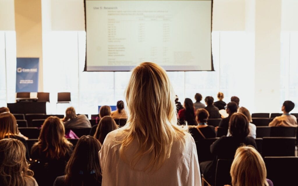 Imagem de uma apresentação em evento, onde uma mulher de cabelos longos está assistindo a uma palestra focada em desenvolvimento profissional.