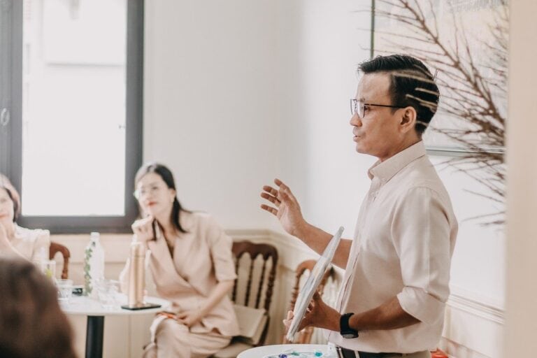 Um homem em pé apresenta uma palestra para um grupo de mulheres sentadas em um ambiente iluminado e moderno. O palestrante segura uma paleta de cores e gesticula enquanto fala, transmitindo informações em um clima de aprendizado.