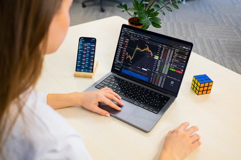 Pessoa analisando gráficos de trading em um laptop enquanto utiliza um smartphone para acompanhar o mercado financeiro.
