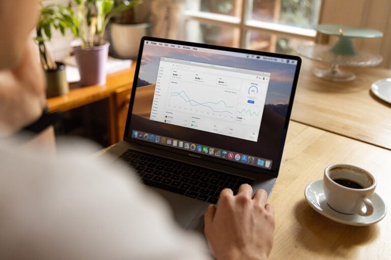 Pessoa usando laptop com gráficos financeiros na tela, acompanhando dados de desempenho. Xícara de café e ambiente aconchegante.